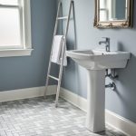 Bright small bathroom interior in an older home with updated chrome faucet and space-saving sink, light gray/white porcelain tile flooring, and soft daylight through a window.